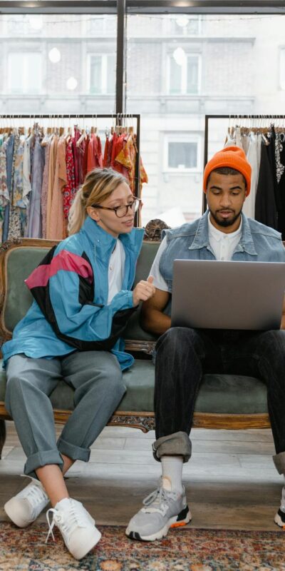 A couple sits on a couch in a stylish clothing store, browsing a laptop together.